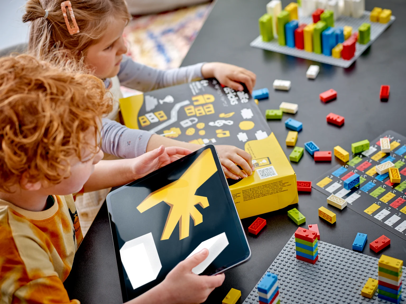 箱の点字に触れる子とタブレットの遊びの案内画面を見る子。机に色の違う点字ブロックが並ぶ